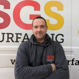 Joe, a white man with dark brown hair, is standing with his arms crossed in front of an SGS Surfacing van. He is wearing a dark grey hoodie with the SGS logo embroidered in red, orange and yellow, and looking directly at the camera.